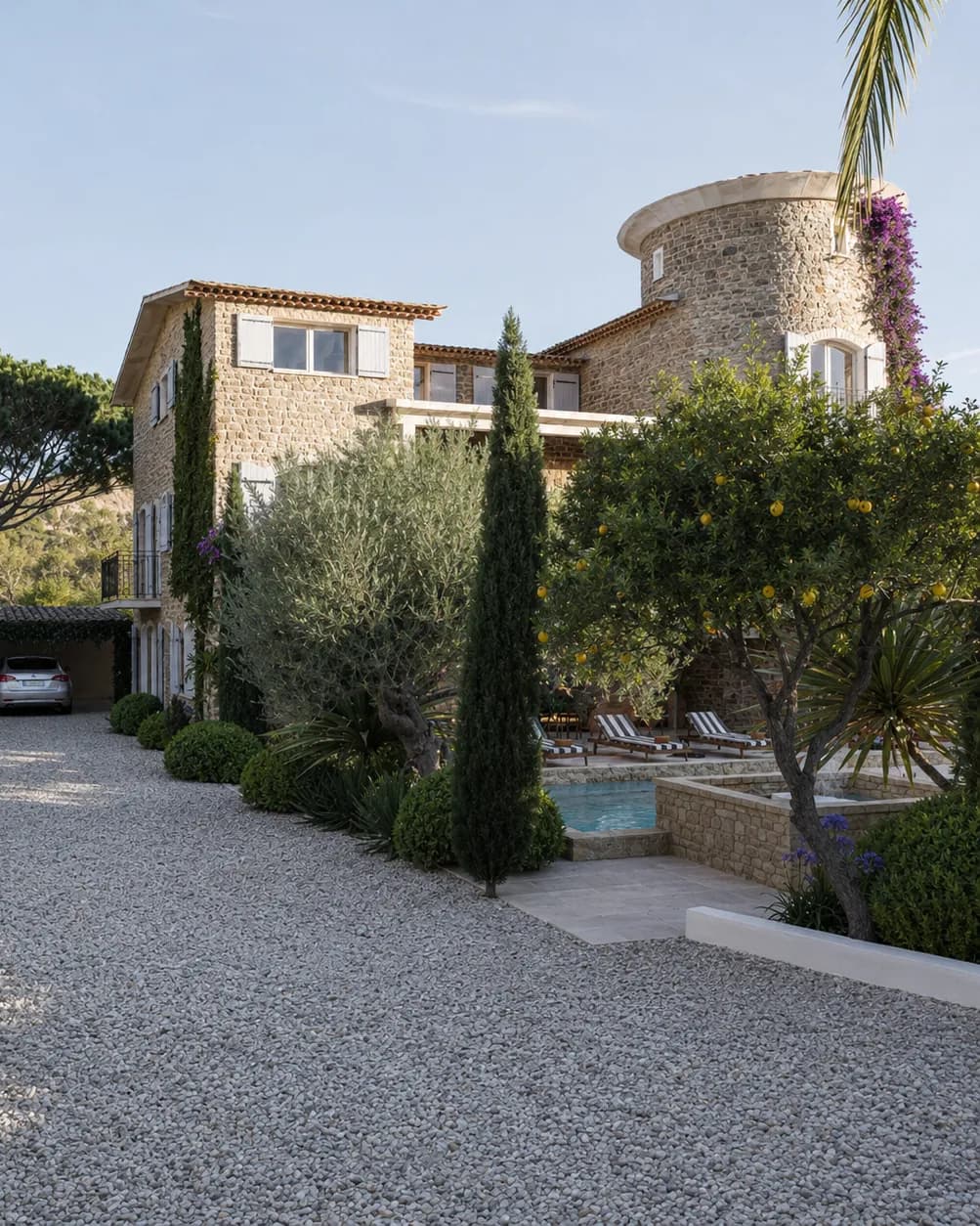 Bastide en pierre avec tourelle ronde, cour de gravier clair bordée de cyprès, oliviers et agrumes, piscine en contrebas derrière un muret de pierre